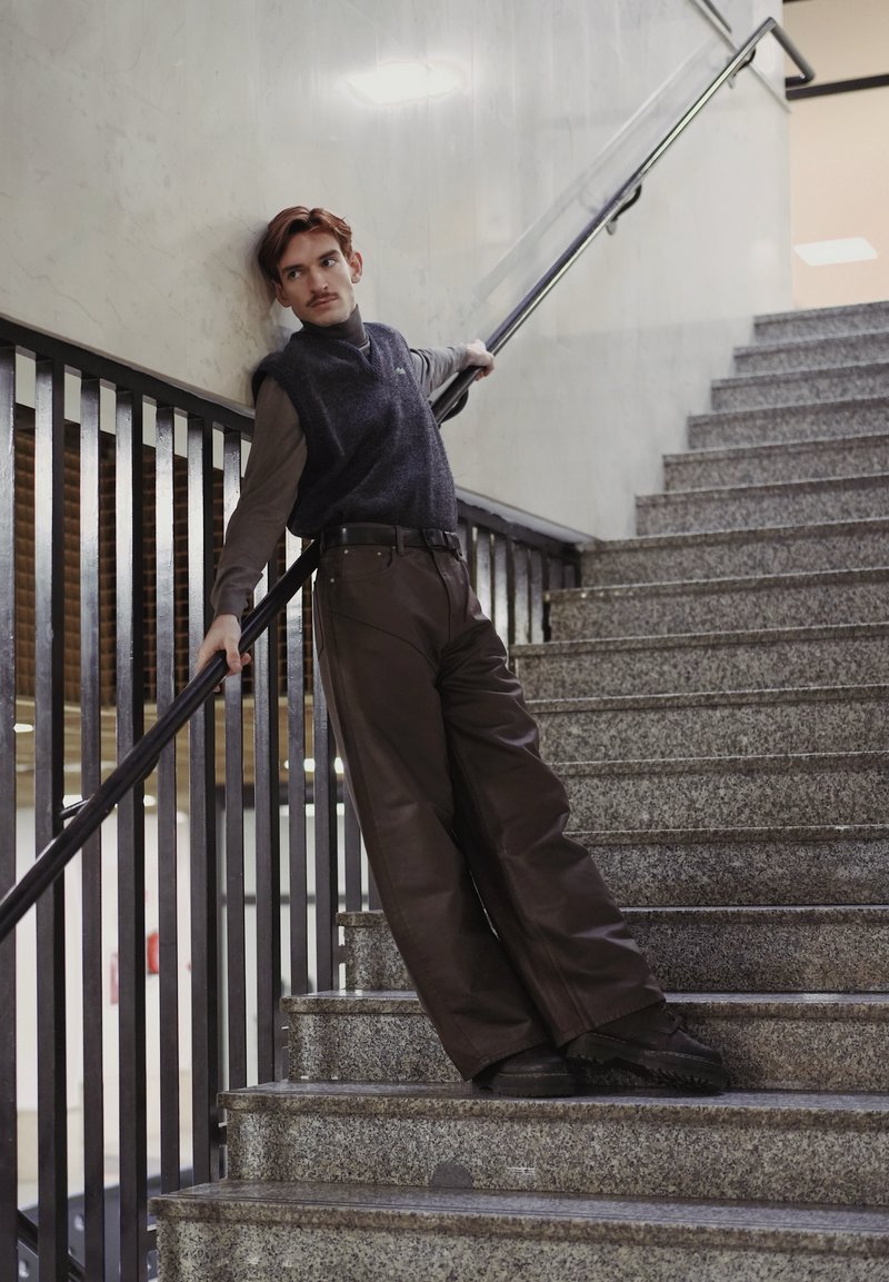 Jeune homme appuyé contre la rampe d'escalier, portant un pantalon foncé et un pull sans manches, regardant sur le côté sur un escalier en pierre à l'intérieur.