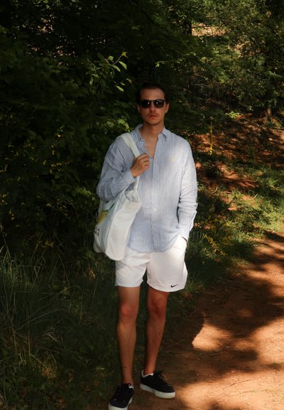 Camisa de botones a rayas azul y blanca, pantalones cortos blancos, zapatillas negras y una bolsa de lona de color claro. Entorno natural con vegetación.