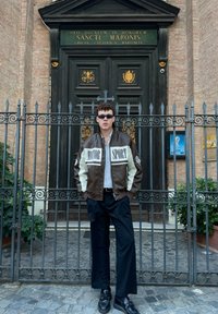 Hnedo-krémová kožená bunda s farebnými blokmi a nápisom "MOTOR SPORT"; čierne široké nohavice; čierne topánky; slnečné okuliare; stojí pri kovanom vchode.