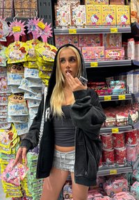 Black oversized hoodie, gray crop top, and light gray ruffled shorts. Holding pink packaging. Colorful merchandise displayed in background.