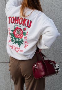 Λευκό φούτερ με κόκκινο και πράσινο λουλουδάτο σχέδιο και κείμενο "TOHOKU PREMIUM". Καφέ παντελόνι και μια μικρή μπορντό τσάντα με charm.