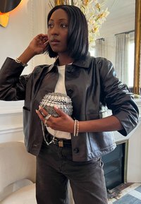 Schwarze Lederjacke, weißes T-Shirt und graue Hose mit einer metallischen Kettenhandtasche. Mehrere silberne Ringe und ein Perlenarmband am Handgelenk.