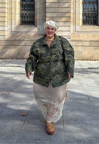 Jachetă camuflată peste o fustă din satin bej cu dantelă; pantofi platform maro; beretă deschisă cu logo. Fundal urban cu arhitectură din piatră.