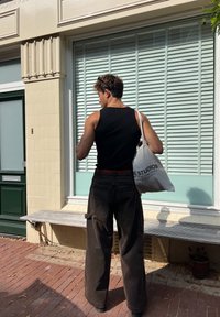 Svart ribbestrikket tanktop, løse mørke cargo-bukser med lommer, hvit tote-bag med teksten "STUDIOS", står ved et vindu med grønn ramme.