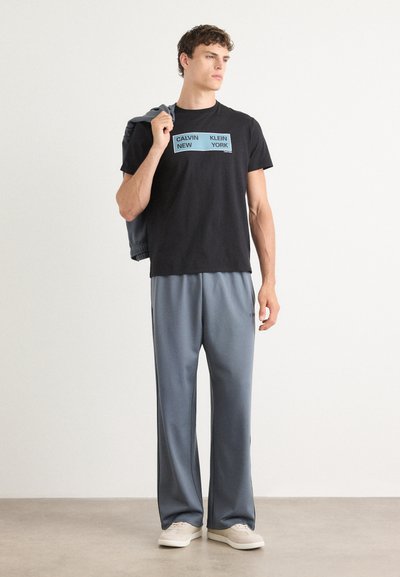 Camiseta negra con texto azul "CALVIN NEW KLEIN YORK", combinada con pantalones deportivos holgados gris claro y zapatillas beige.