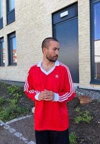 Camisola Adidas de manga longa vermelha com gola e detalhes em mangas brancos, apresentando riscas verticais e logotipo. Usada sobre calças pretas.