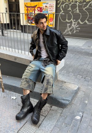Joven con chaqueta de cuero negra, camisa blanca y pantalones cortos camuflaje rasgados sentado en un bordillo en una calle urbana con graffiti y carteles detrás.