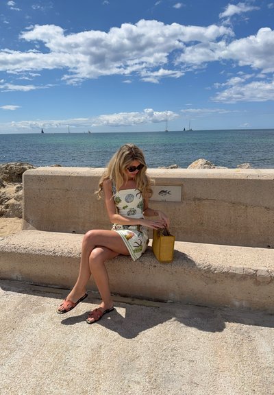 Vestido con estampado floral y escote cuadrado, sentada en un banco de piedra. Sosteniendo un bolso tejido amarillo, usando gafas de sol negras y sandalias marrones.
