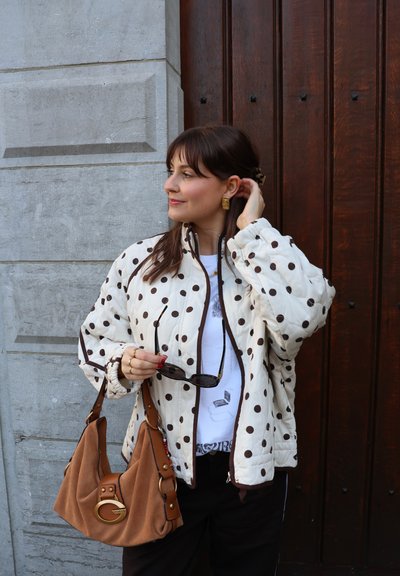 Mujer de pie junto a una puerta de madera, vestida con una chaqueta blanca con lunares, sosteniendo gafas de sol y un bolso marrón, mirando hacia un lado.