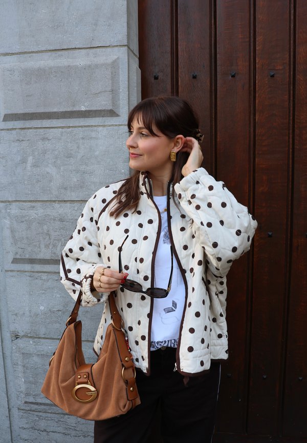 Kvinne står ved en tredør, iført en hvit jakke med prikker, holder solbriller og en brun håndveske, ser mot siden.