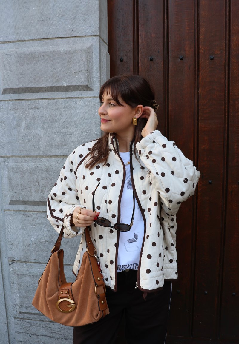 Kvinde står ved en trædør iført en hvid jakke med prikker, holder solbriller og en brun håndtaske, og kigger til siden.