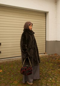 Manteau en fourrure noire avec un motif texturé et un col haut, associé à un sac à main floral. Une longue jupe grise complète la tenue.