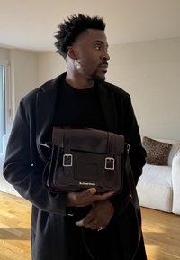 Homme portant un manteau noir et une boucle d'oreille en perle, tenant une sacoche Dr. Martens marron foncé dans un salon baigné de lumière.