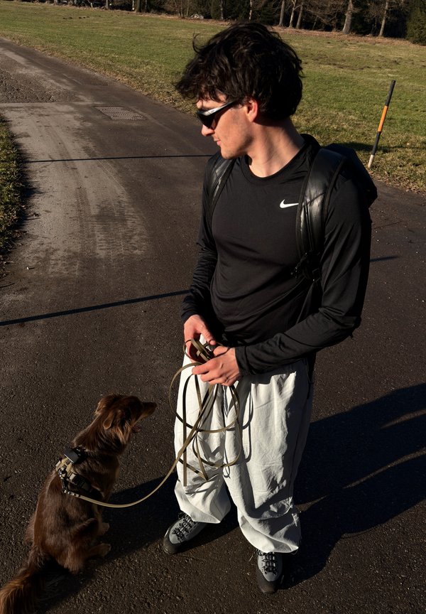 En person i en svart langermet skjorte og hvite bukser holder en hundebånd, med en brun hund som sitter i nærheten med sele på.