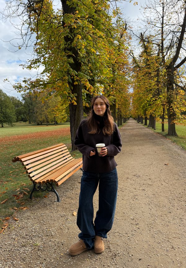Una mujer sostiene un café para llevar en un sendero arbolado de un parque en otoño, junto a un banco de madera.