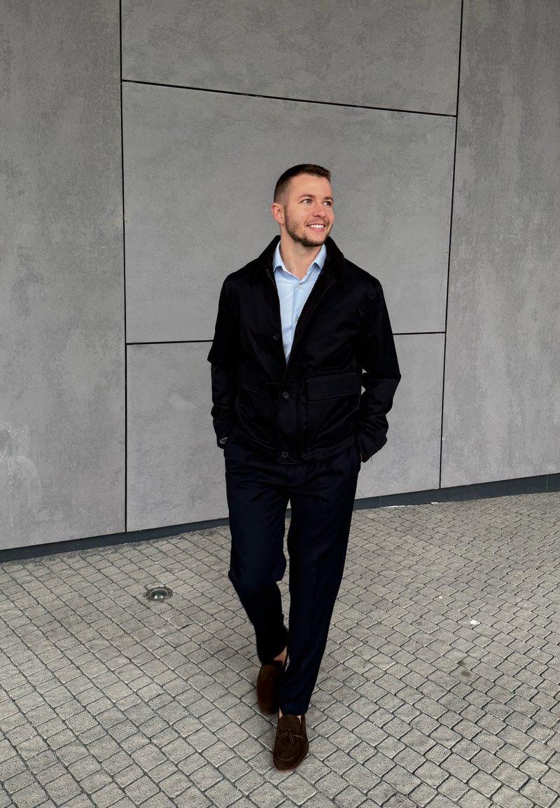 Young man in black jacket and navy trousers walking on tiled pavement, smiling and looking up against a grey paneled wall background.