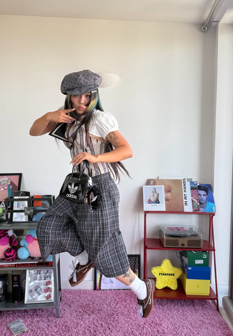 Young person wearing a gray polka-dot beret jumps indoors, holding a black Adidas bag, with plaid pants and brown shoes on a pink carpet.