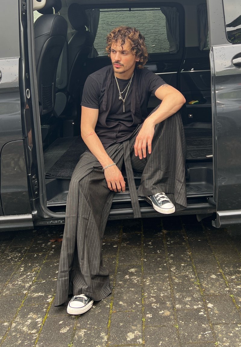 Jeune homme aux cheveux bouclés assis sur la marche d'un van, portant un t-shirt noir, un pantalon large à fines rayures et des baskets noires, regardant sur le côté.