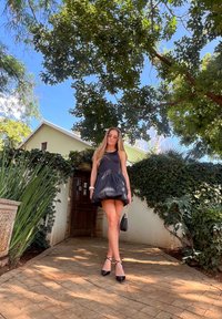 Μαύρο αμάνικο φόρεμα με φούστα που συγκεντρώνεται, γυαλιστερή υφή. Συνδυασμένο με μαύρες ψηλές γόβες και μια μικρή τσάντα, μπροστά σε πράσινο φόντο.