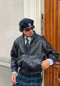Chaqueta bomber de cuero negro con cierre de cremallera, de ajuste holgado, llevada sobre una camisa blanca y pantalones de cuadros. Un boina negra y gafas de sol complementan el conjunto.