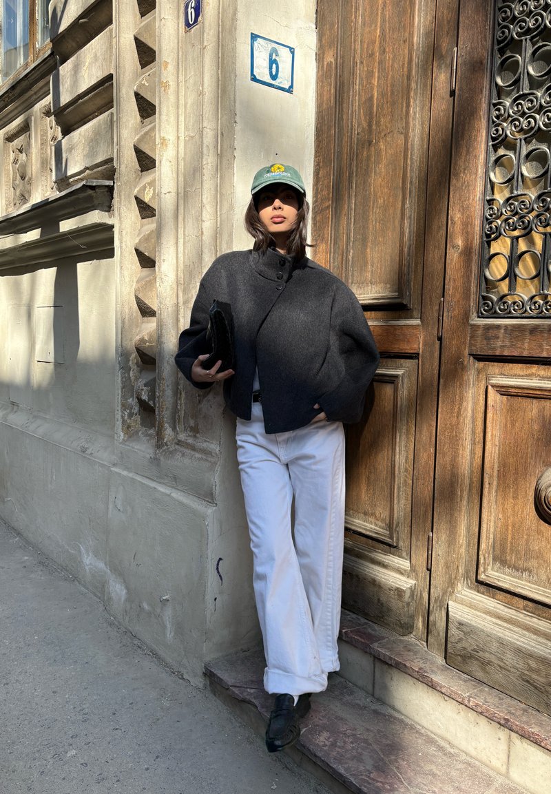 Femme portant une casquette verte, une veste sombre et un pantalon blanc, appuyée contre un bâtiment en pierre près de portes en bois numérotées 6.