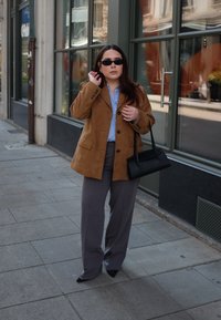 Bruine suède blazer boven een lichtblauwe button-up, grijze wijde broek en een zwarte handtas. Zonnebril maakt de look compleet. Stedelijke setting.