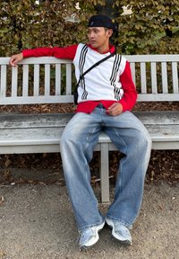 Κόκκινο μπλουζάκι με μακρύ μανίκι κάτω από λευκή αμάνικη μπλούζα με ρίγες. Χαλαρά, ξεθωριασμένα μπλε τζιν. Γκρίζα αθλητικά παπούτσια. Καθισμένος σε ξύλινο παγκάκι.