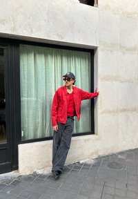 Κόκκινο ελαφρύ μπουφάν πάνω από ένα κόκκινο πουκάμισο, συνδυασμένο με χάρη σκούρους γκρι παντελόνες και μαύρα παπούτσια. Σταθείτε μπροστά σε ένα παράθυρο με πράσινες κουρτίνες.