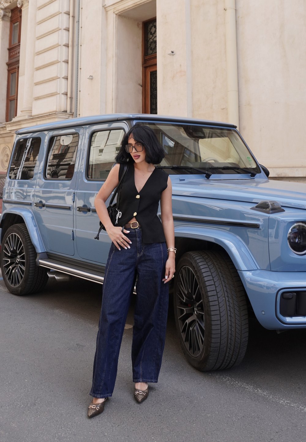 SUV bleu garé dans la rue. Le modèle porte un haut sans manches noir avec des boutons, un jean large en denim foncé et des chaussures à motifs, tenant un sac.