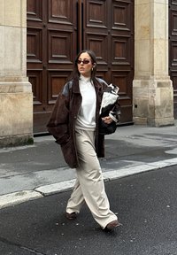 Brun oversized jakke med sorte læderærmer, hvid rullekrave, beige bukser med vide ben og brune sko. Holder en sort og hvid taske.