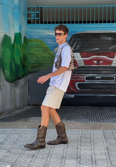 Una persona vestida con una camiseta blanca, pantalones cortos beige y botas de vaquero marrones, caminando con un bolso de piel marrón texturizado sobre su hombro.