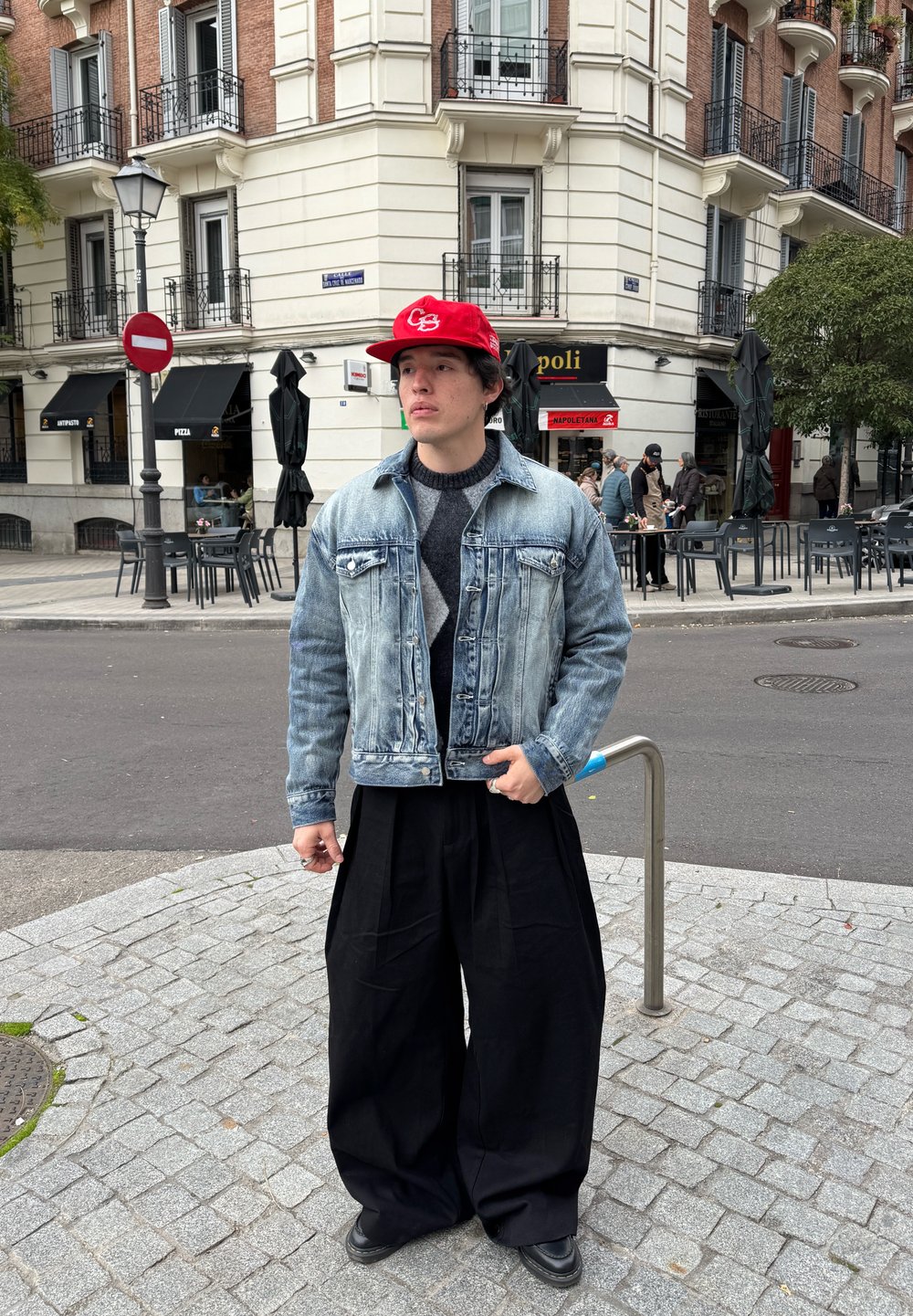 Jeune homme portant une casquette rouge, une veste en denim et un pantalon large noir se tenant à un coin de rue pavée dans une ville européenne.