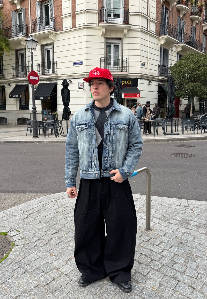 Young man wearing a red cap, denim jacket, and wide black pants standing on a cobblestone street corner in a European city.