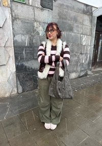 Jeune femme aux cheveux courts, pull rayé, gilet en fourrure, pantalon large, tenant un sac gris, debout sur un trottoir mouillé contre un mur en pierre.