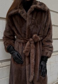 Manteau en fausse fourrure marron avec une taille ceinturée, un grand col et deux boutons. Associé à des gants en cuir noir. Texture douce, apparence luxueuse.