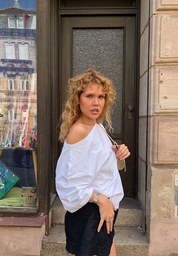 Junge Frau mit lockigem Haar, die ein weißes Schulter-freies Oberteil und einen schwarzen Rock trägt, steht an einer dunklen Tür und Steinstufen in urbaner Umgebung.