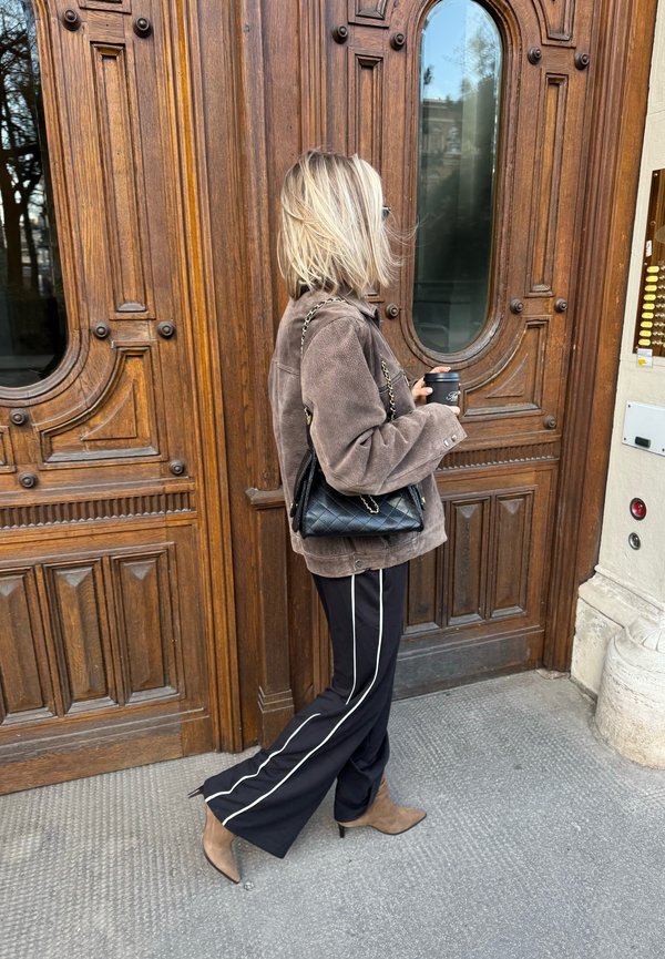 Woman with blonde hair walking past a large ornate wooden door, holding a coffee cup, wearing brown jacket, black pants with white stripes, and brown boots.