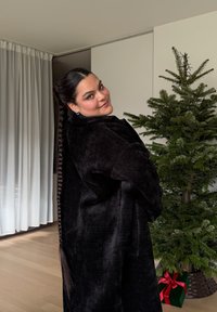 Sort, tekstureret oversize frakke båret over en top med høj krave. Lang fletning og markante øreringe. Grønne træer og gaver i baggrunden.