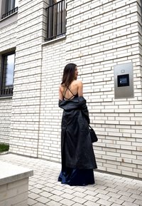 Femme en manteau noir en cuir par-dessus une robe bleu marine, tenant un sac à main, debout près d'un mur de briques blanches avec un panneau interphone.