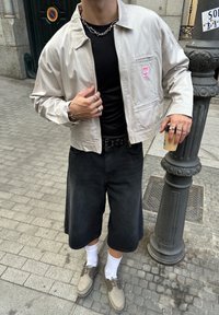 Gaiši pelēka zip-up jaka ar vertikālām svītrām, melna T-kreklis, brīvi melni šorti, pelēki apavi, balti zeķes, sudraba ķēde un gredzeni.