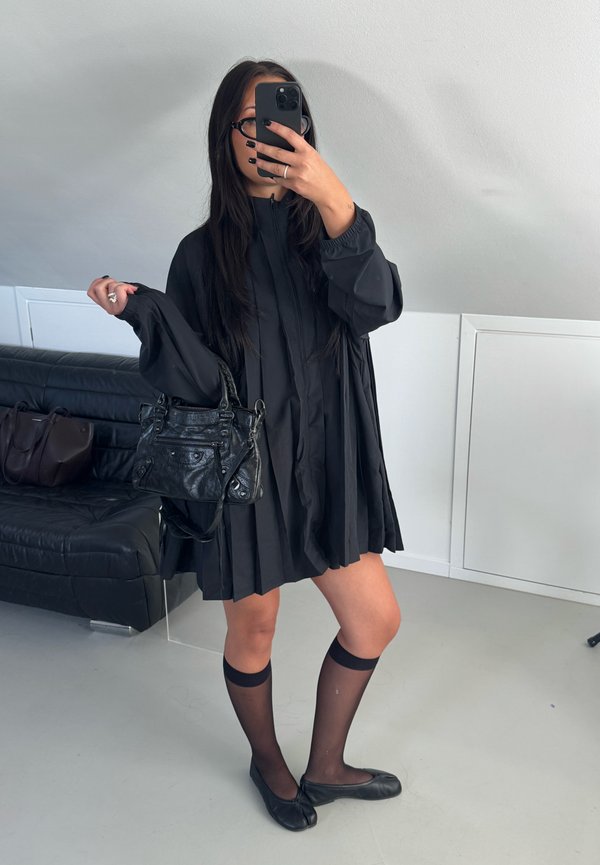 Woman wearing black pleated dress, sheer knee-high socks, and black flats, holding a black handbag, taking a mirror selfie indoors.
