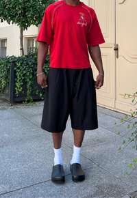 Κόκκινο κοντομάνικο πουκάμισο με γραφικά και κείμενο, μαύρες φαρδιές βερμούδες, λευκές κάλτσες και μαύρες σαμπό; στέκεται πάνω σε μια πέτρινη επιφάνεια με πράσινους θάμνους.