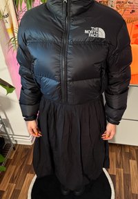 Črna puffer jakna z visokim ovratnikom in zadrgo; vsebuje logo The North Face. Slojevita črna krilo s plissiranjem in teksturirano tkanino.