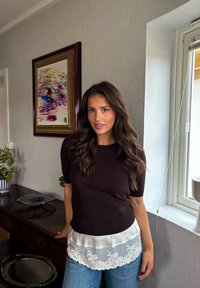 Joven con cabello largo y ondulado, vestida con una blusa negra con dobladillo de encaje y jeans azules, de pie junto a un aparador de madera cerca de una ventana y una pintura abstracta.