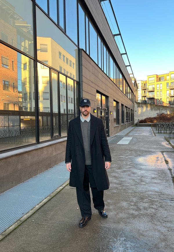 Osoba u dugom crnom kaputu, tamnim hlačama i crnom šeširu stoji na vlažnoj stazi pored reflektirajućih prozora moderne zgrade.