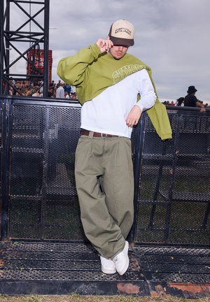 Grüner und weißer übergroßer Pullover, der über einem weißen Mesh-Oberteil getragen wird, kombiniert mit lockeren olivgrünen Hosen und weißen Turnschuhen. Beigefarbene Kappe mit Logo.