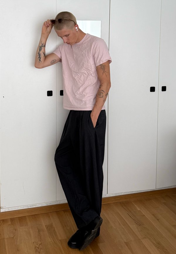 Jeune personne aux cheveux courts et tatouages porte des lunettes de soleil, des écouteurs, un t-shirt rose clair et un pantalon noir ample, debout à l'intérieur sur un sol en bois.
