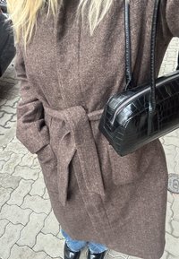 Brown herringbone coat with a tie belt, paired with a small black crocodile-patterned handbag and black shiny shoes.