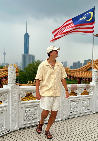 Camisa corta de manga corta color beige claro con patrones texturizados, pantalones cortos blancos y sandalias marrones; el fondo presenta edificios de gran altura y una bandera.