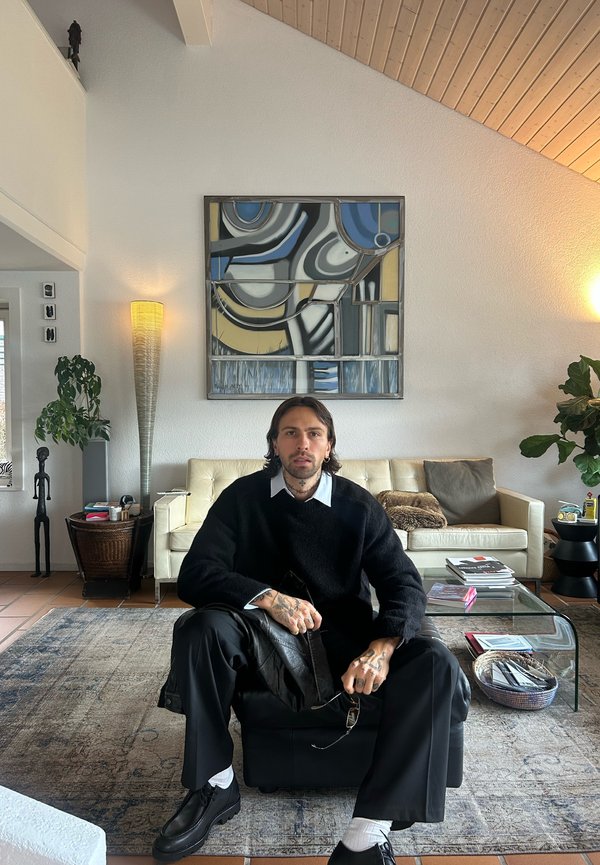 Svart strikkegenser, hvit skjorte med krage, svarte bukser og sko. Sitter på en svart puff ved et glassbord, omgitt av dekor.